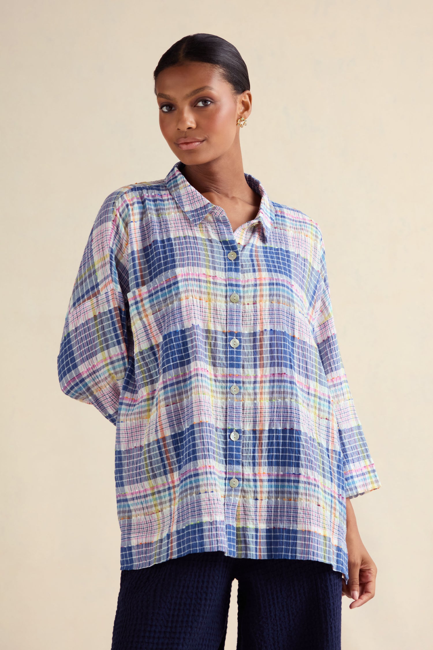 A woman stands against a plain background wearing the Crinkle Check Jacquard Shirt, a loose-fitting long-sleeve top in blue, yellow, pink, and green tones.