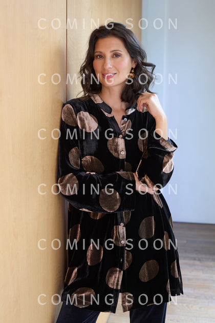A woman with shoulder-length dark hair models the Silk Velvet Spot Shirt—a versatile black top with large beige polka dots—while standing against a wooden wall. "COMING SOON" is repeatedly overlaid on the image.