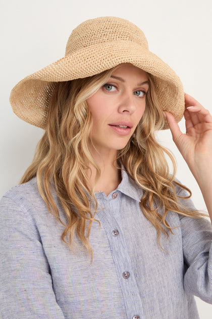 A woman with long blonde hair models the Handwoven Raffia Tie Hat and a light blue button-up shirt, looking at the camera against a plain background.