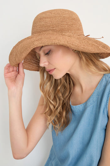 Woman wearing a sleeveless blue top tilts her head down, touching the brim of her Handwoven Raffia Tie Hat.