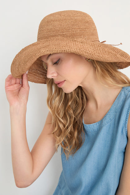 Woman wearing a sleeveless blue top tilts her head down, touching the brim of her Handwoven Raffia Tie Hat.