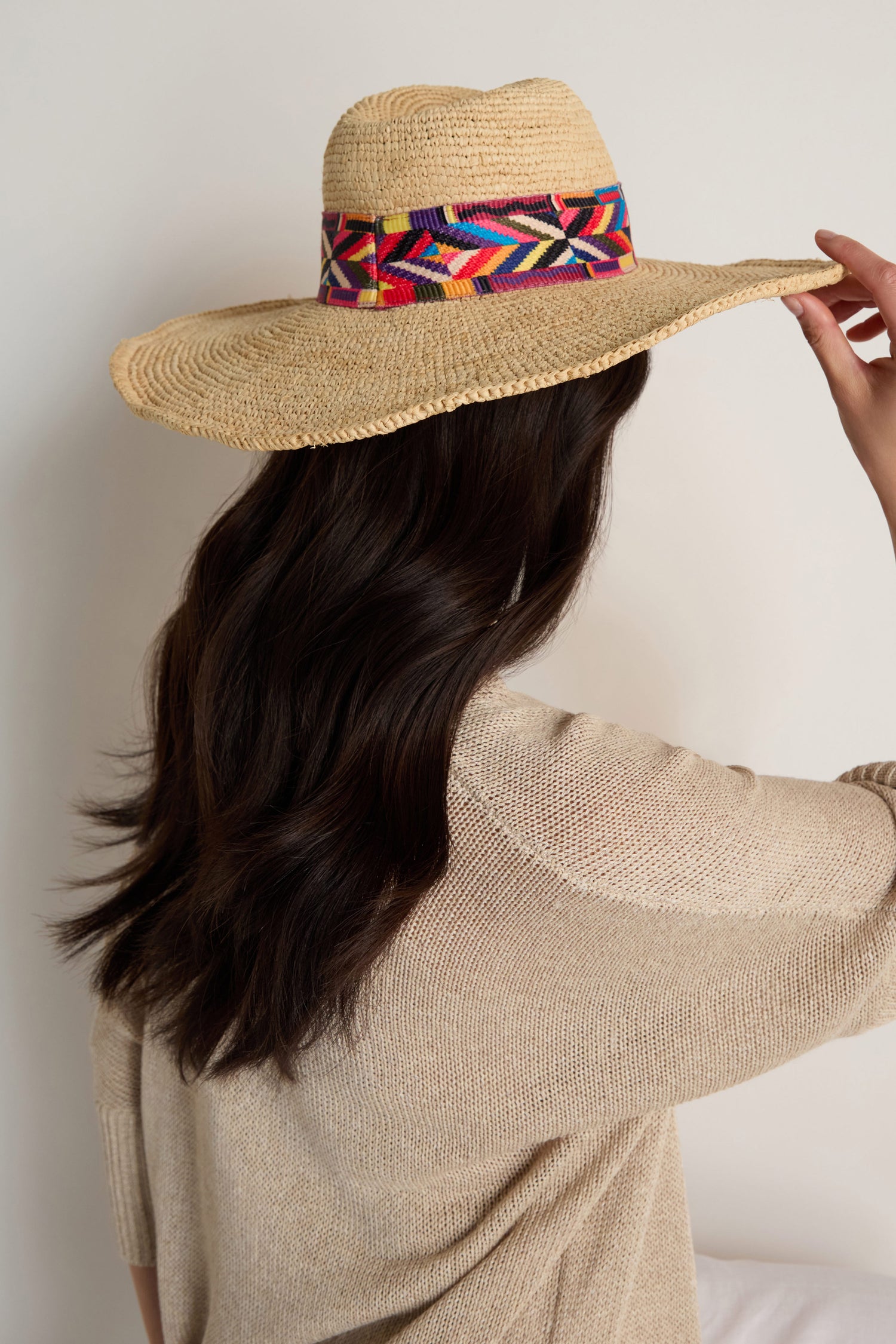 A person with long dark hair in a beige sweater wears the Handwoven Raffia Western Hat, featuring a colorful band, seen from behind against a plain background to showcase its artisanal style and sun protection.