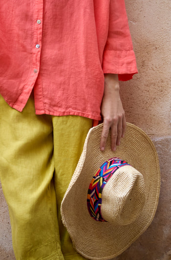 A person in a coral shirt and yellow pants holds the Handwoven Raffia Western Hat, featuring a colorful band, while standing against a textured beige wall in the TON1160B finish.