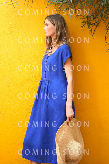 A woman in a blue dress stands against a bright yellow wall, holding the Handwoven Raffia Wide Brim Hat. The code TON1176 is subtly displayed as “COMING SOON” text repeats over the image.
