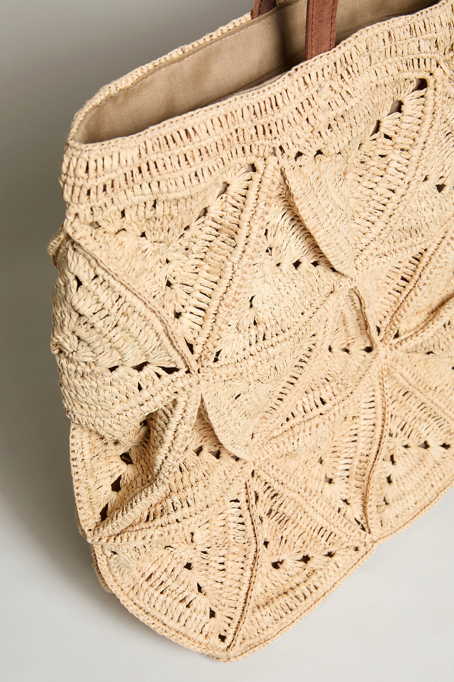 The Handwoven Raffia Basket Bag, inspired by Madagascar baskets, showcases a beige geometric petal crochet design with brown handles and is photographed against a neutral background.