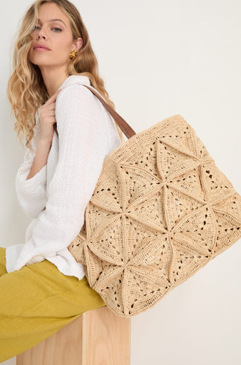 A woman with wavy blonde hair, in a white blouse and yellow pants, sits on a wooden block carrying the Handwoven Raffia Basket Bag—a large beige tote with a geometric pattern—over her shoulder.
