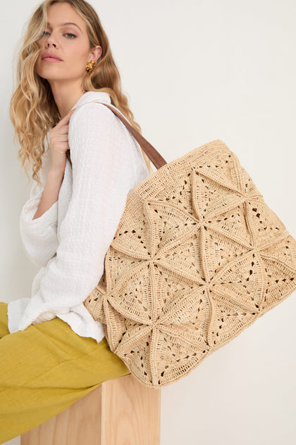 A woman with wavy blonde hair, in a white blouse and yellow pants, sits on a wooden block carrying the Handwoven Raffia Basket Bag—a large beige tote with a geometric pattern—over her shoulder.