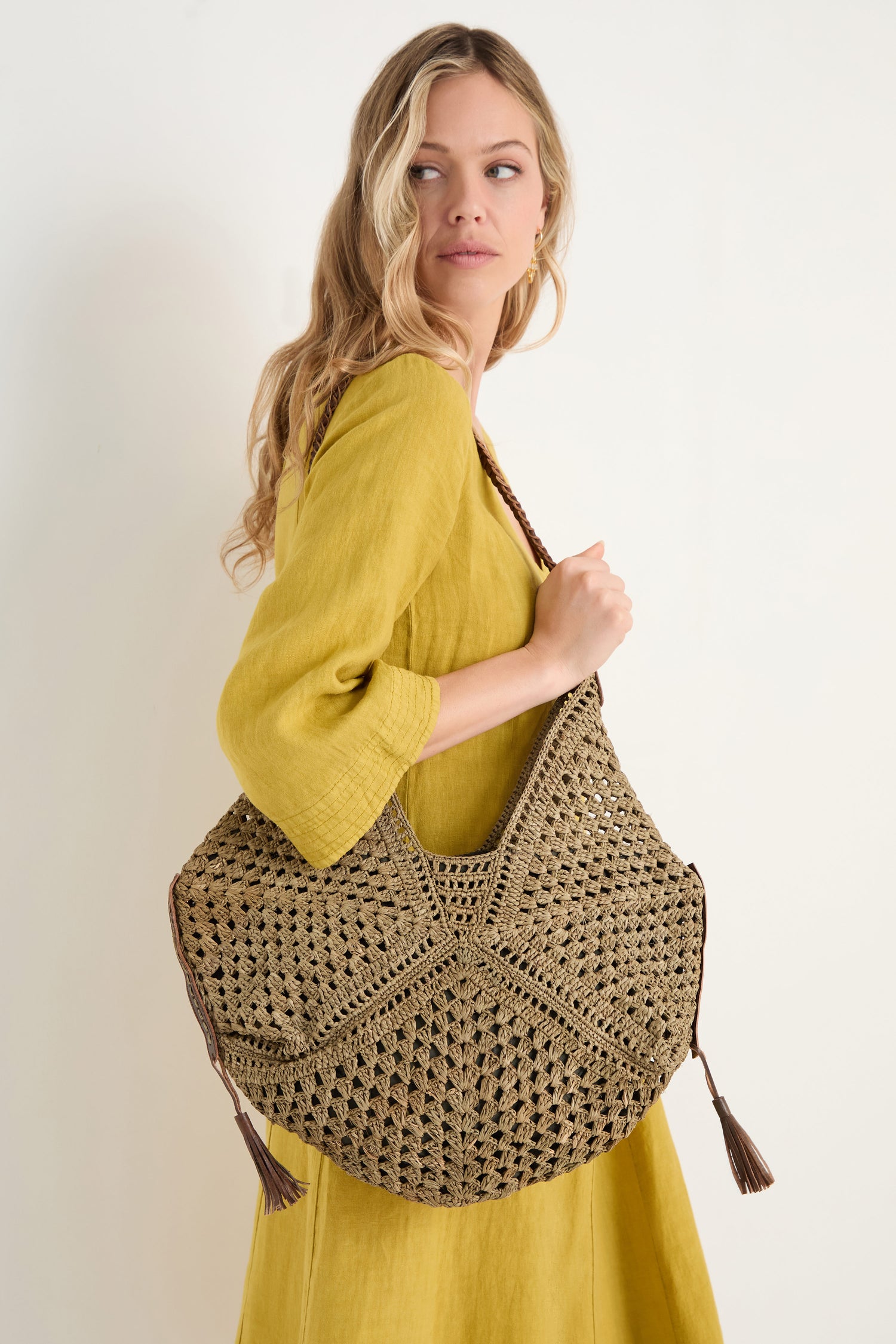 A woman in a yellow dress stands sideways against a plain background, carrying the Handwoven Raffia Tassel Detail Handbag over her shoulder.