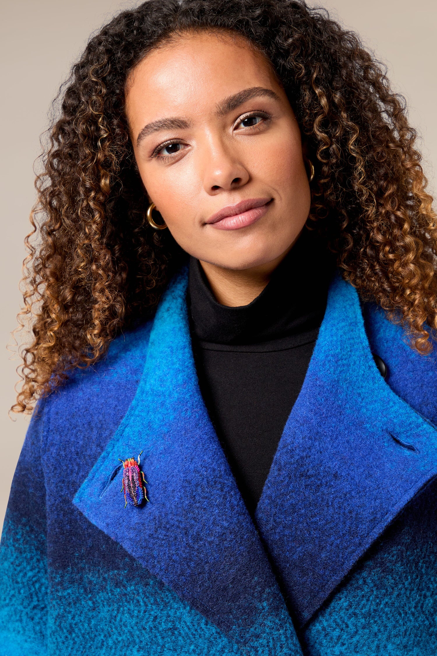 A woman wearing a blue and black coat with a Rainbow Leaf Beetle Brooch that adds a touch of sparkle.