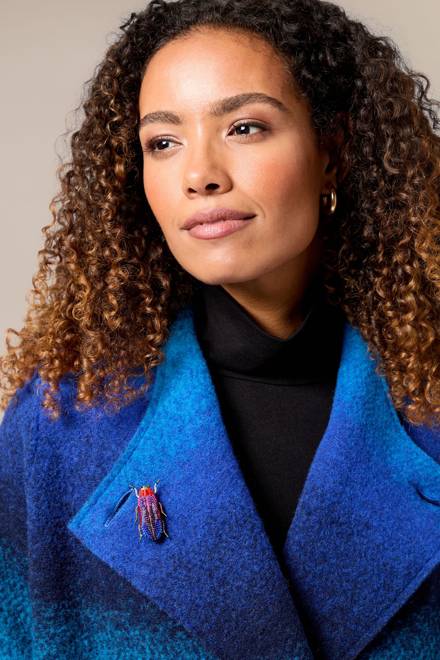 A woman in a blue coat with curly hair, wearing a Rainbow Leaf Beetle brooch.