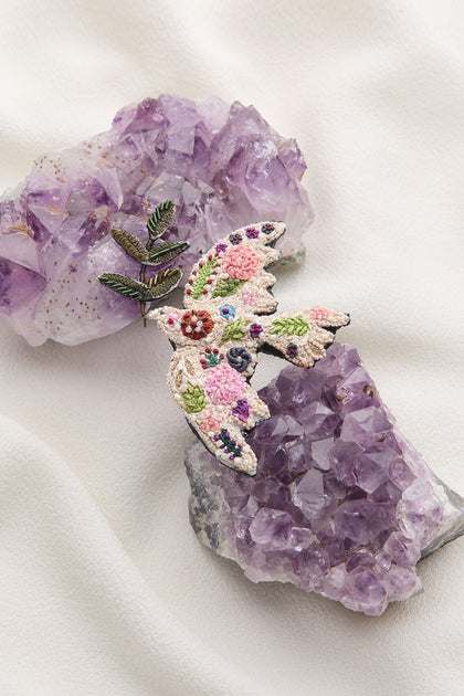 The Hand Embroidered Peace Dove Brooch showcases a Peace Dove holding a green leafy twig, perched on a cluster of purple amethyst crystals against a white fabric background.