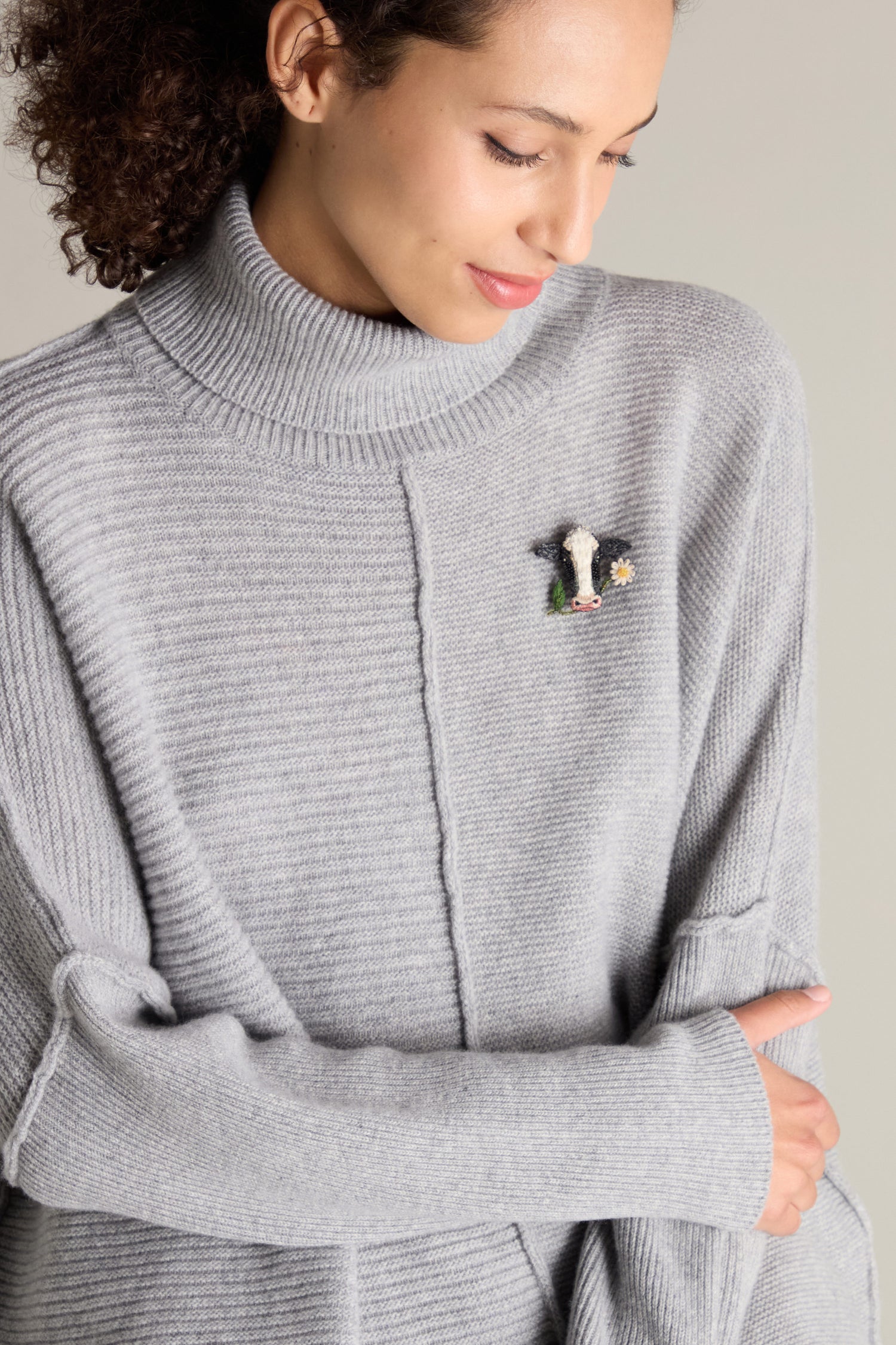 A person with curly hair wears a gray ribbed turtleneck sweater adorned with a Hand Embroidered Cow Brooch.