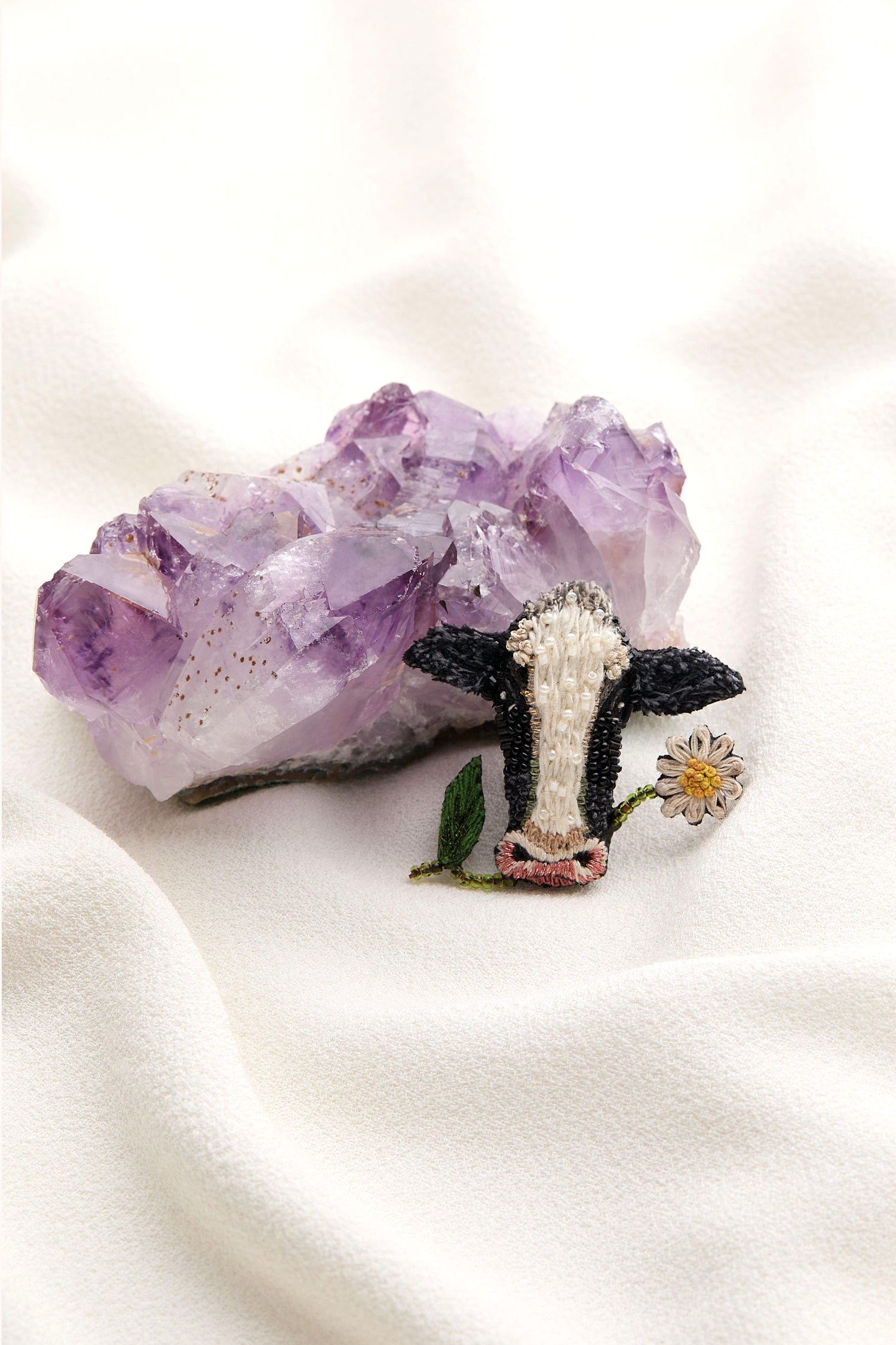 A whimsical touch is added by placing the Hand Embroidered Cow Brooch with intricate sequin and beaded detailing and a small white flower beside a large purple crystal on soft white fabric.