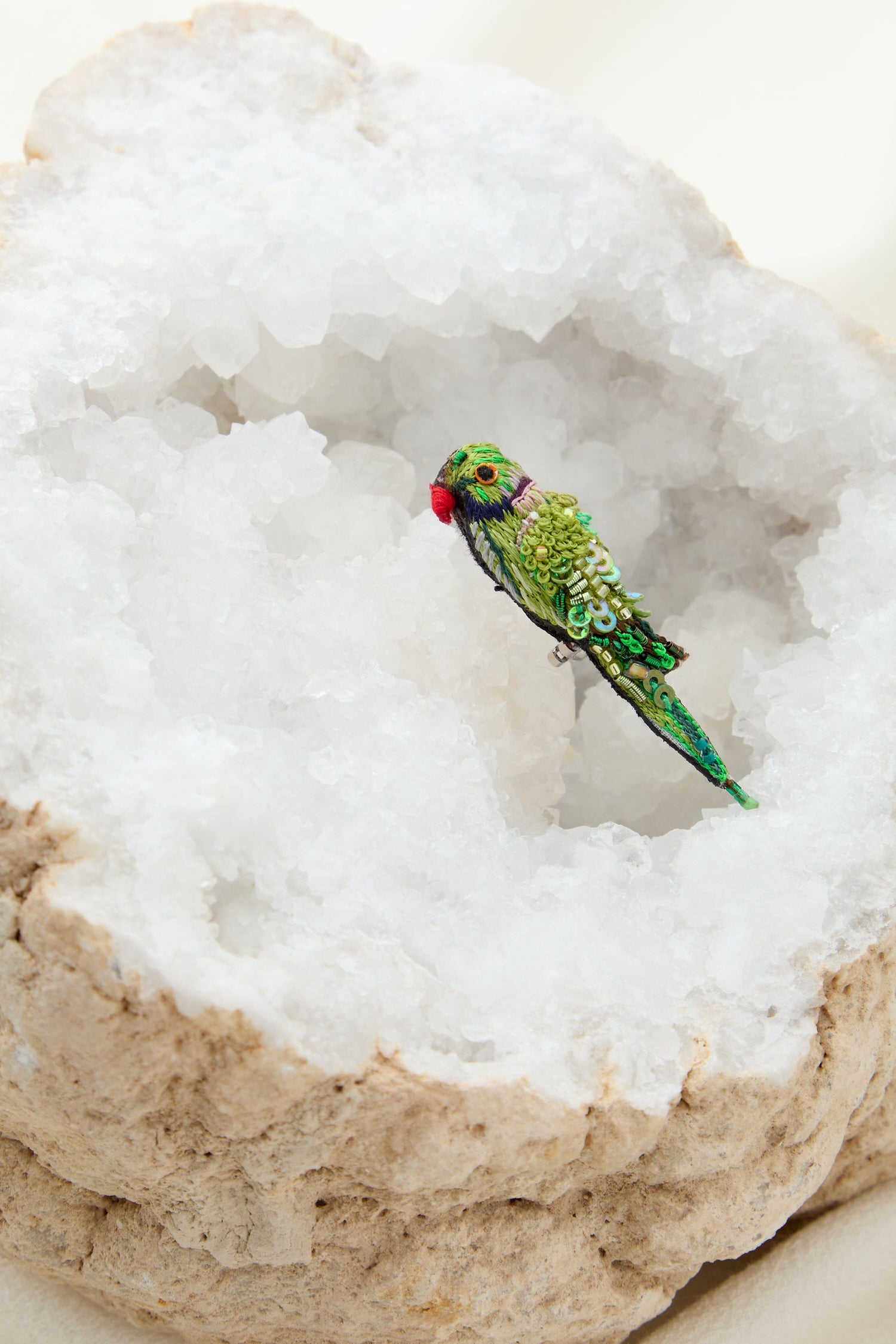 A Hand Embroidered Parrot Brooch sitting on a rock, its plumage sparkling with embroidered beads.