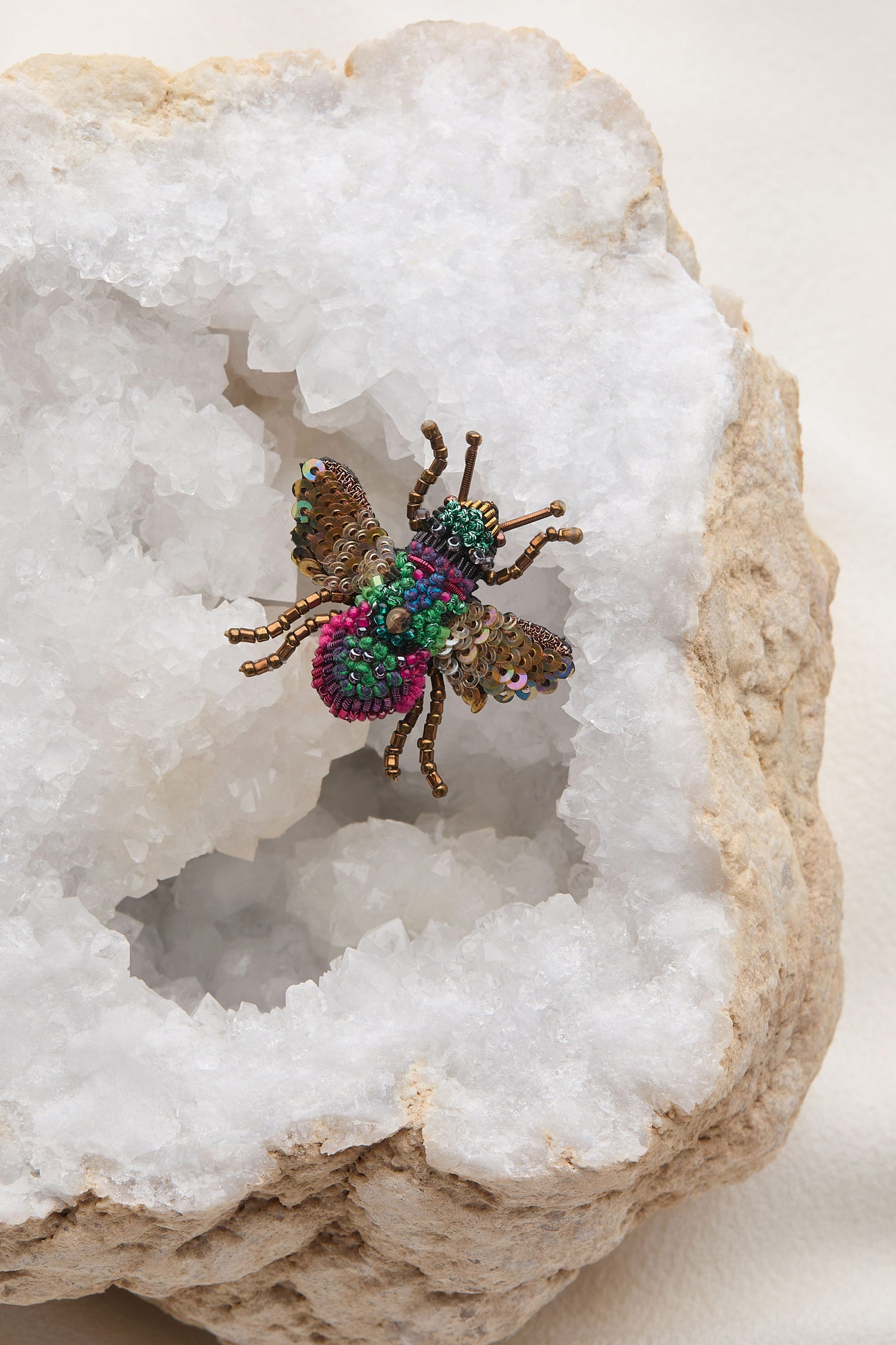 A Hand Beaded Sequin Rainbow Bee Brooch with colorful details is displayed inside a geode with white crystals, perfectly capturing Trovelore's exquisite craftsmanship.