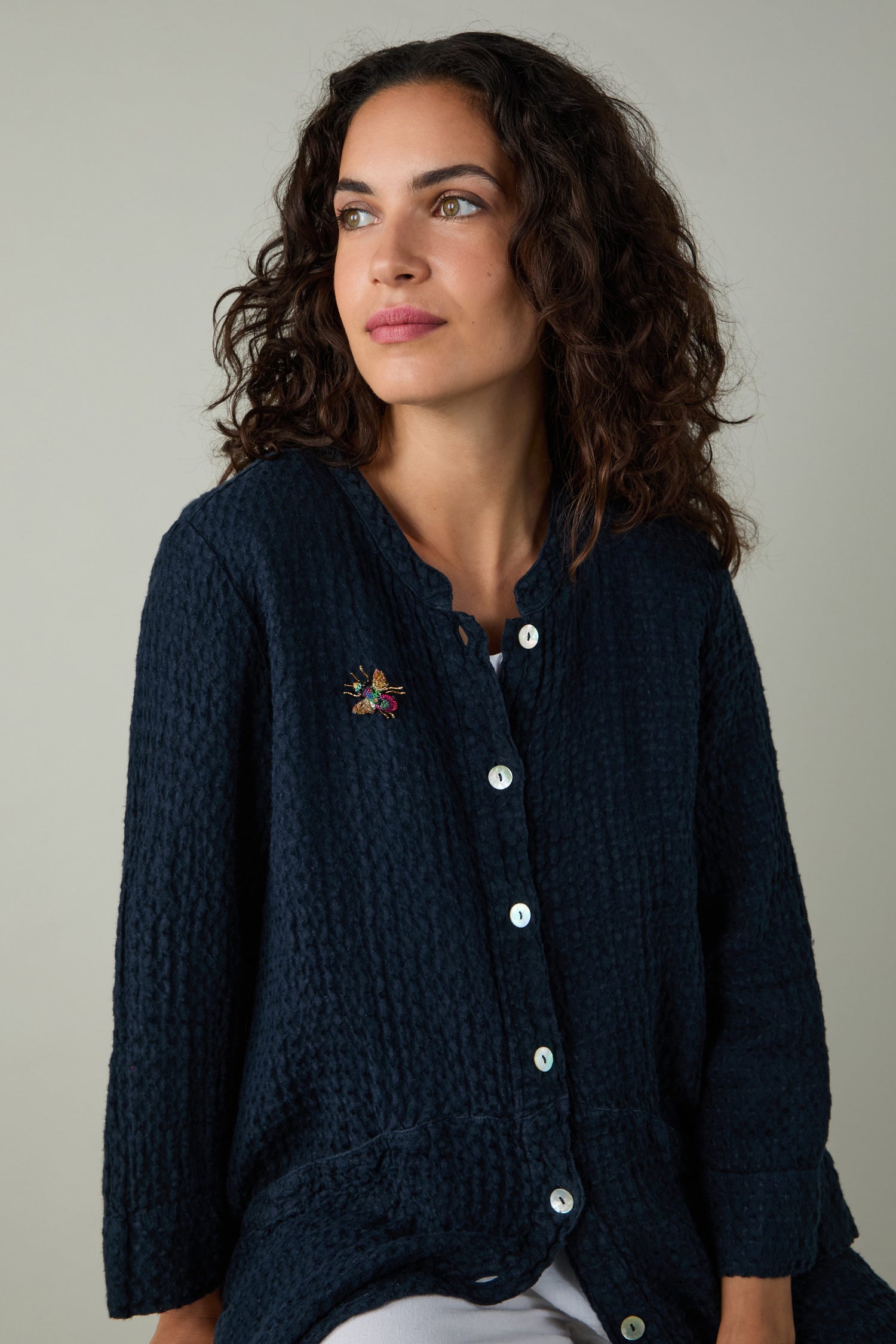 A woman with curly brown hair, dressed in a dark textured button-up shirt, wears the Hand Beaded Sequin Rainbow Bee Brooch and gazes off to the side against a plain background.