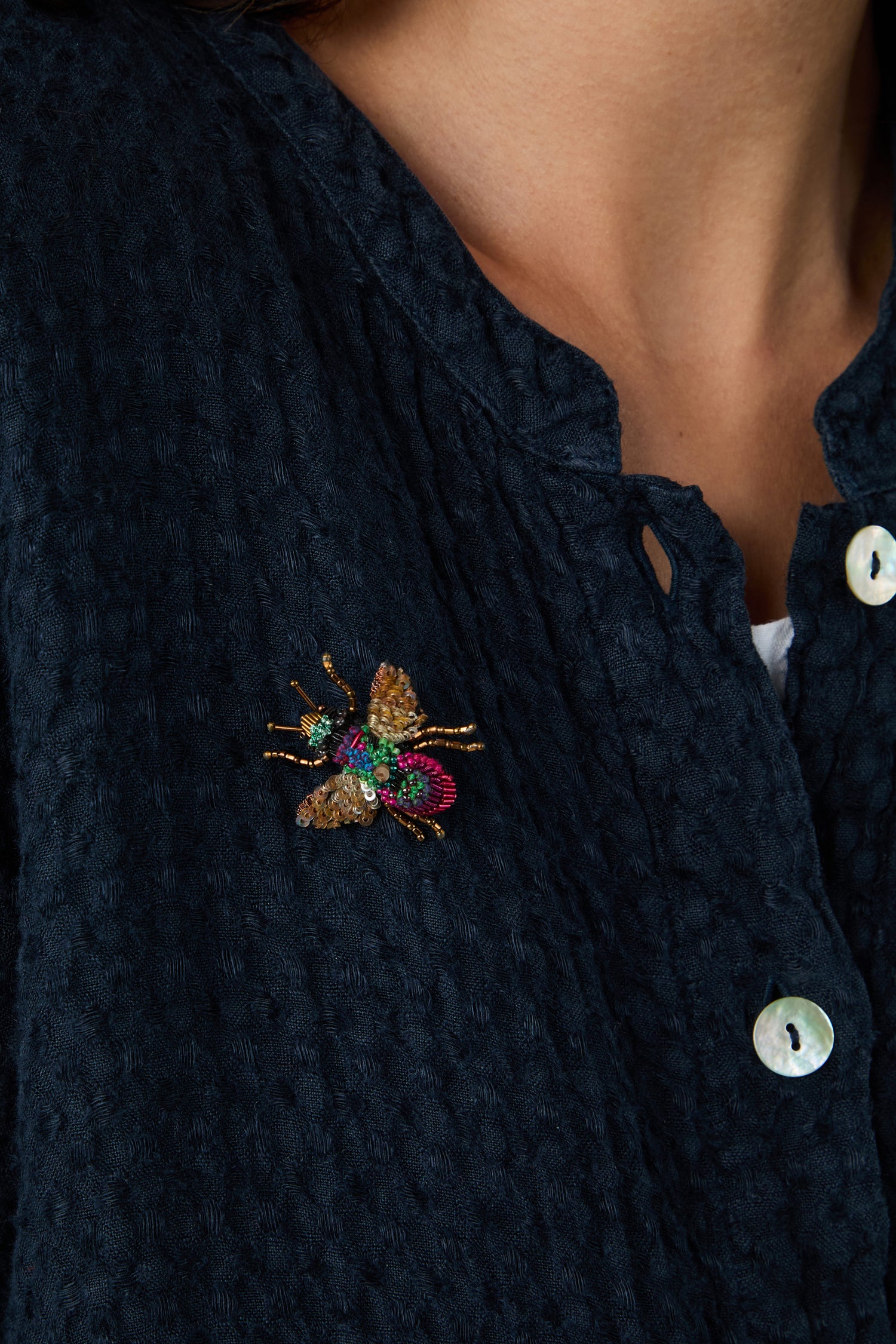 A person wears a dark textured shirt with the Hand Beaded Sequin Rainbow Bee Brooch, adorned with colorful beads, pinned near the left collar.