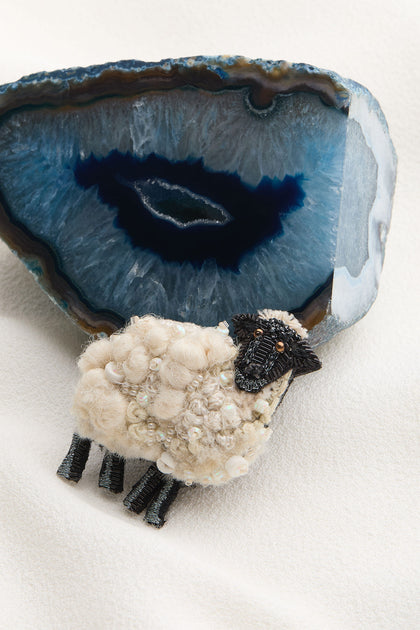 A small sheep figurine made of wool and beads is positioned in front of a large geode with blue and white crystalline layers, echoing the delicate craftsmanship of the Hand Embroidered Ewe Brooch.