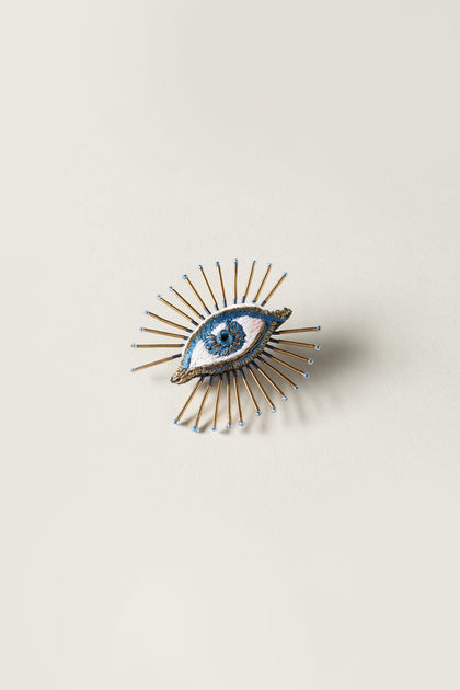 The Hand Embroidered Sea Mystic Eye Brooch features a central blue eye surrounded by gold spikes, all presented on a light background.