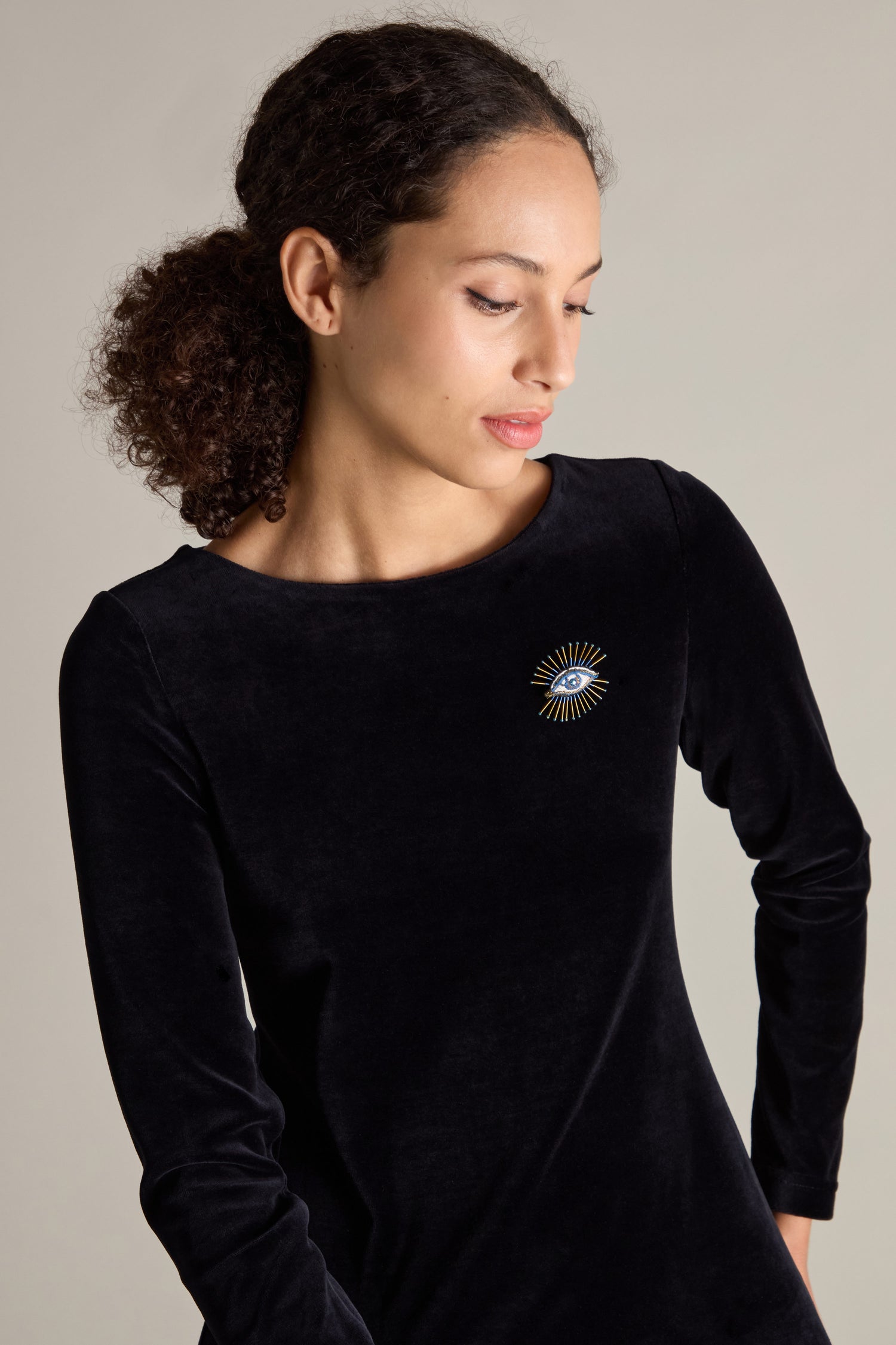 A woman with curly hair wearing a black top adorned with the Hand Embroidered Sea Mystic Eye Brooch on the chest stands against a plain background.