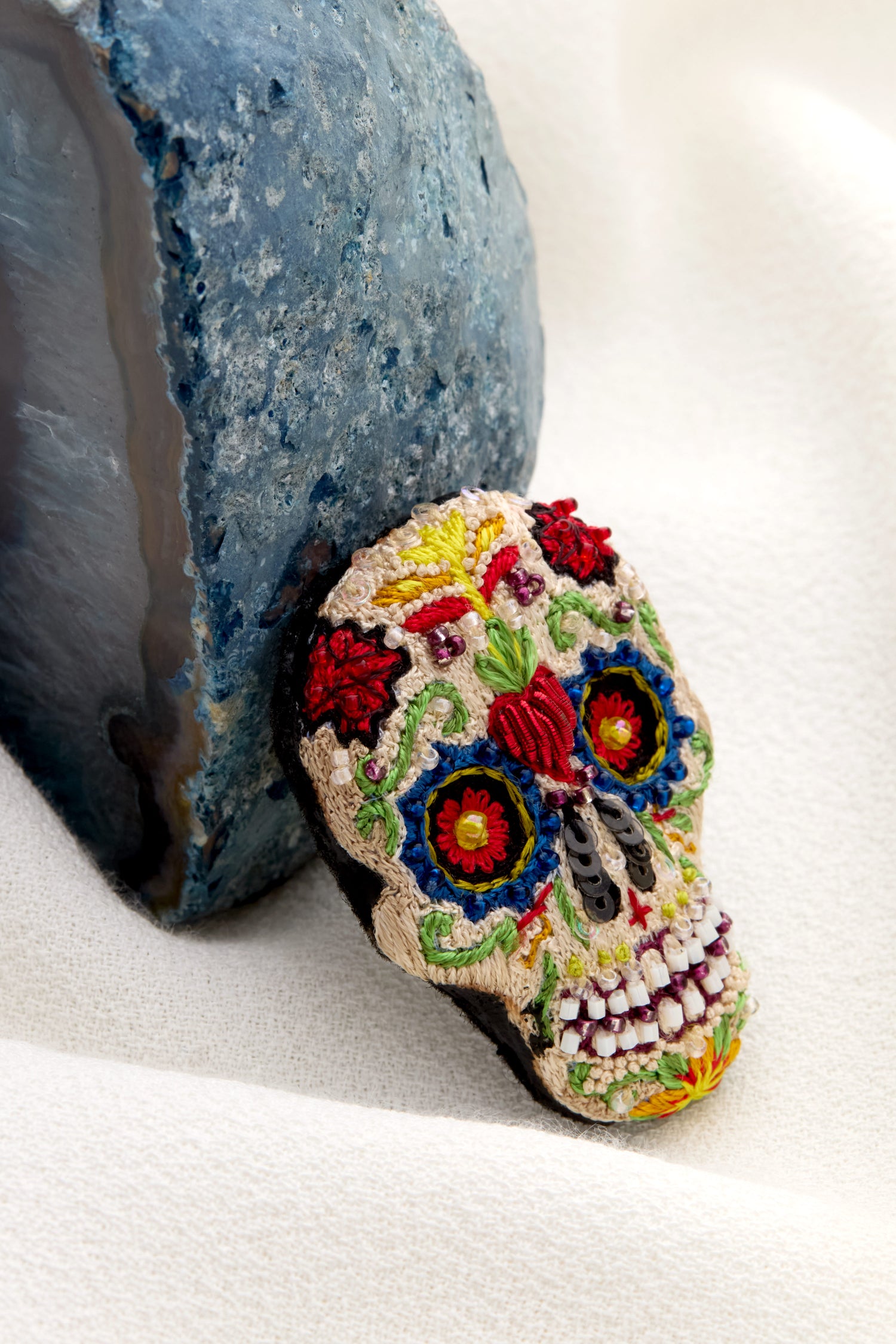 A Hand Embroidered Calavera Brooch, featuring intricate beaded embellishments, is set against a striking blue geode on a white fabric background.