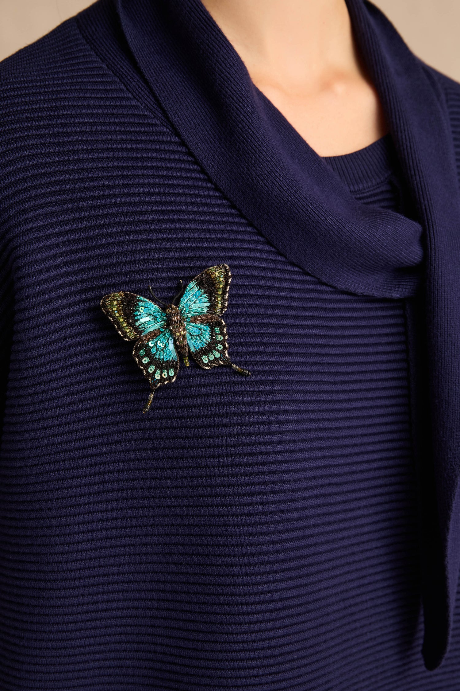 A person wearing a textured, dark blue knit top adorned with a Hand Beaded Sea Green Swallowtail Brooch, featuring shades of turquoise, black, and gold in an exquisite butterfly design.