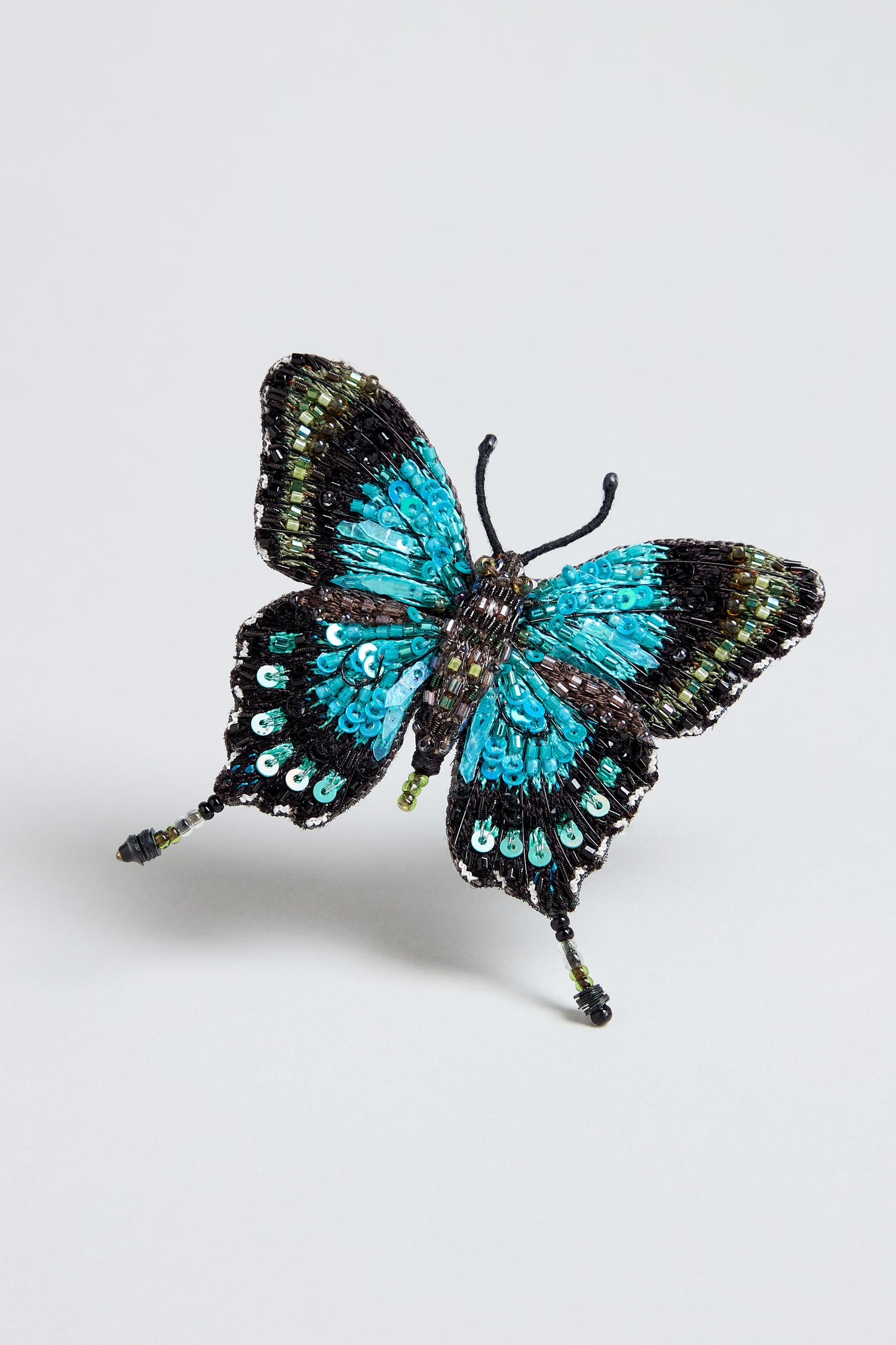 A decorative Hand Beaded Sea Green Swallowtail Brooch with beaded wings, resting gracefully on a light grey surface.