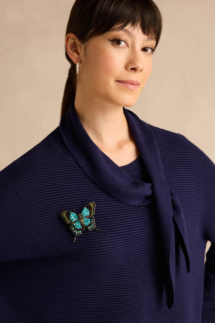 A woman with dark hair wears a blue ribbed sweater adorned with the Hand Beaded Sea Green Swallowtail Brooch, its iridescent sea green and black hand-embroidered details shimmering against a neutral background.