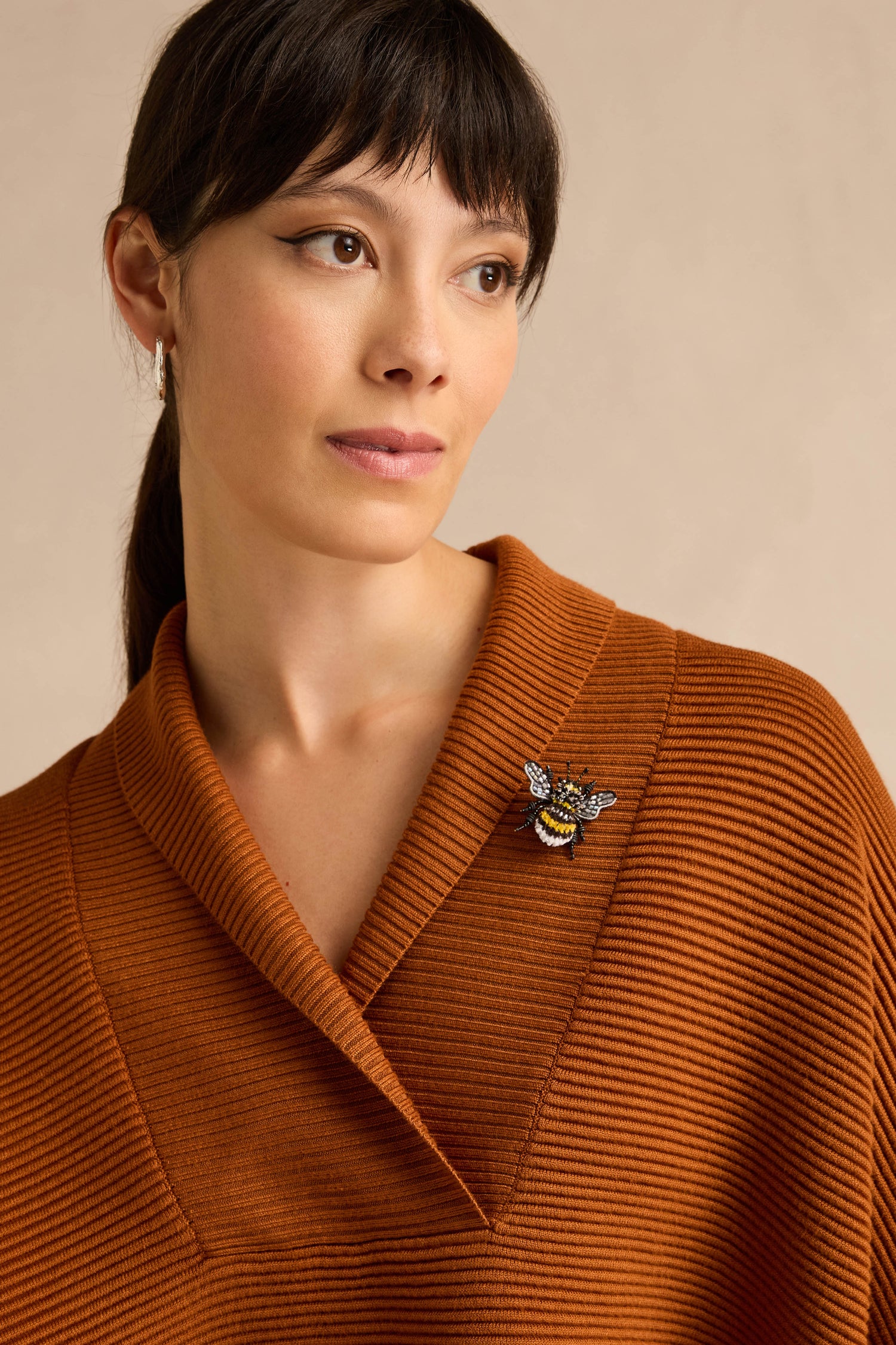 Woman in rust-colored ribbed top wearing a Hand Beaded Yellow-Banded Bee Brooch on the collar, looking slightly to the side with a neutral expression.