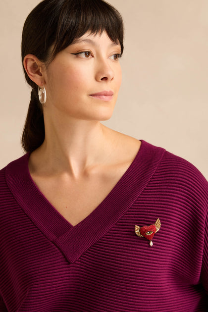 A model with dark hair in a ponytail wears silver hoop earrings and a ribbed purple v-neck sweater, accented with a stunning Hand Beaded Guardian Heart Brooch—an intricately hand beaded accessory featuring delicate wings and a central eye detail.