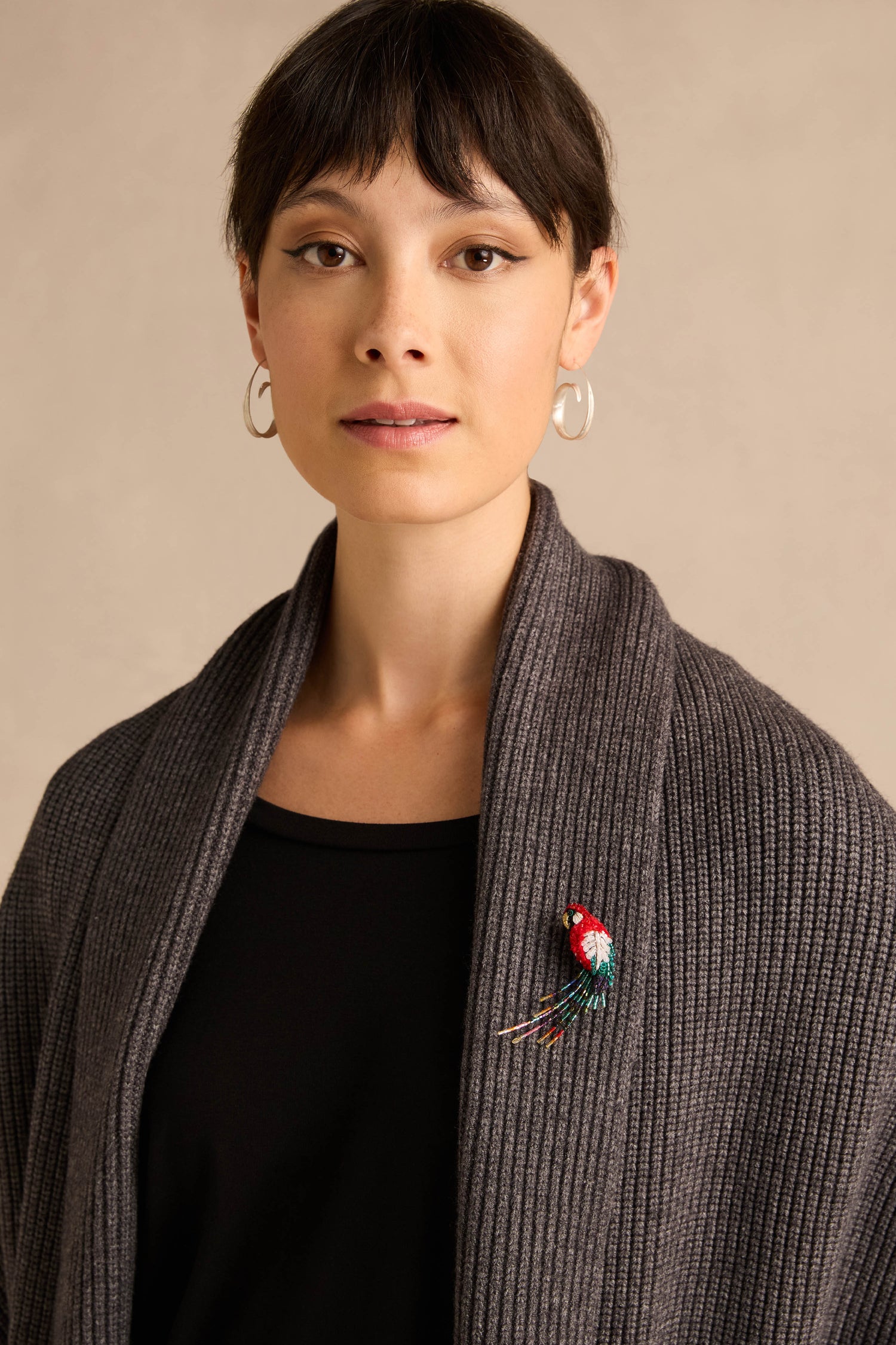 A woman with short dark hair wearing hoop earrings, a ribbed gray shawl, and a black top, has the Hand Beaded Arara Macao Brooch pinned vibrantly to her shawl.