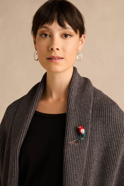 A woman with short dark hair wearing hoop earrings, a ribbed gray shawl, and a black top, has the Hand Beaded Arara Macao Brooch pinned vibrantly to her shawl.