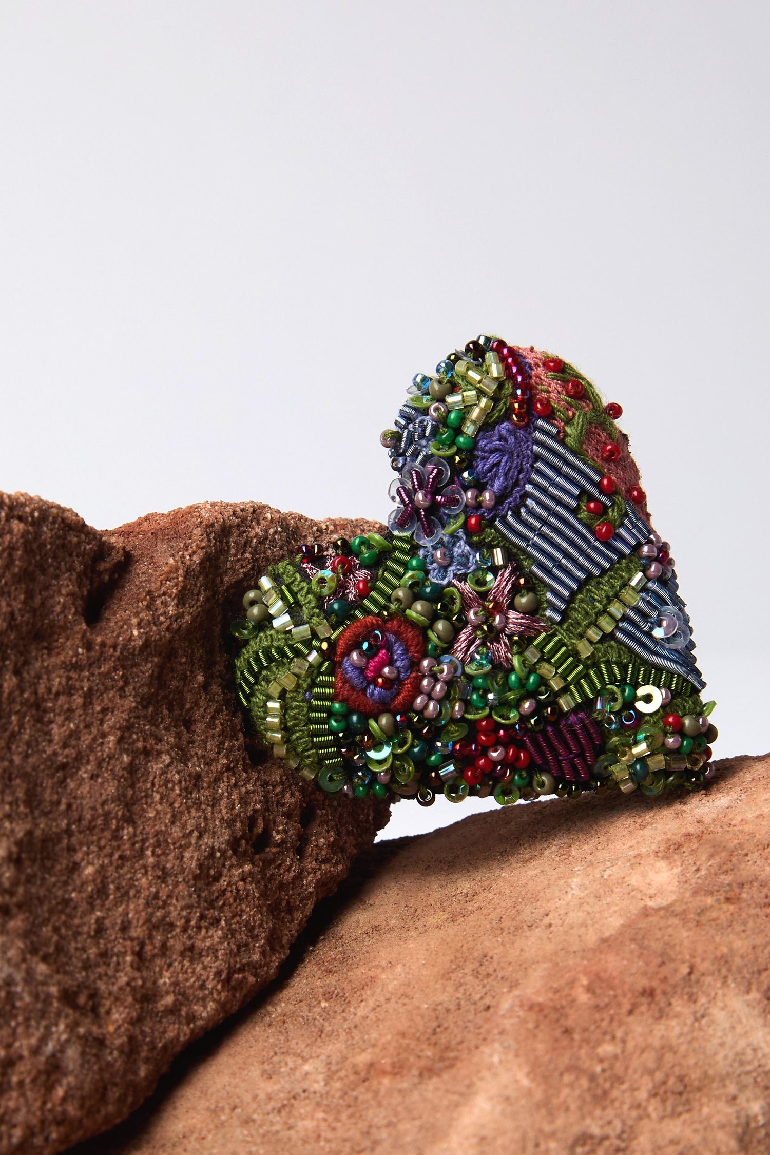 The Hand Beaded Blooming Heart Brooch by Trovelore, showcasing intricate hand embroidery and vibrant beads with floral patterns, elegantly rests between two brown rocks.