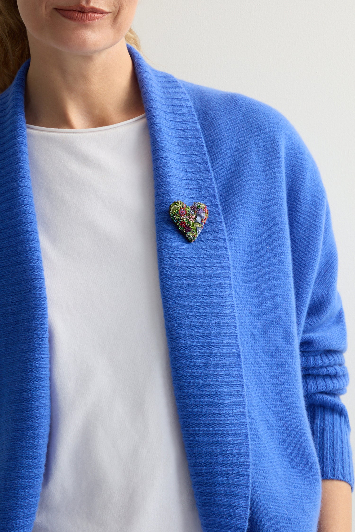 Wearing a blue cardigan over a white shirt, they complete their look with the intricately detailed Hand Beaded Blooming Heart Brooch by Trovelore, adding a touch of elegance.