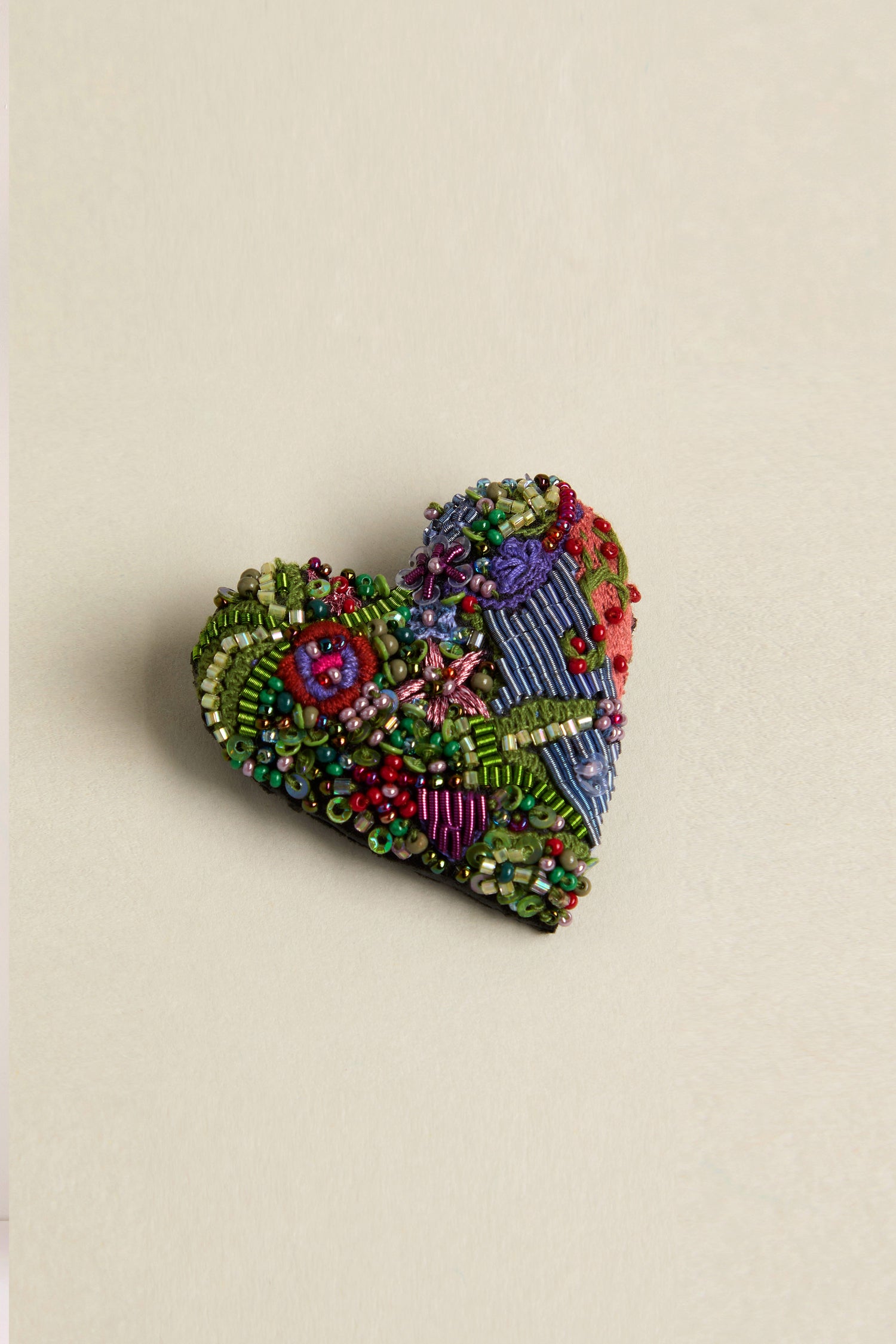 Discover the Hand Beaded Blooming Heart Brooch by Trovelore, a heart-shaped accessory showcasing vibrant hand embroidery and intricate beadwork on a neutral backdrop.