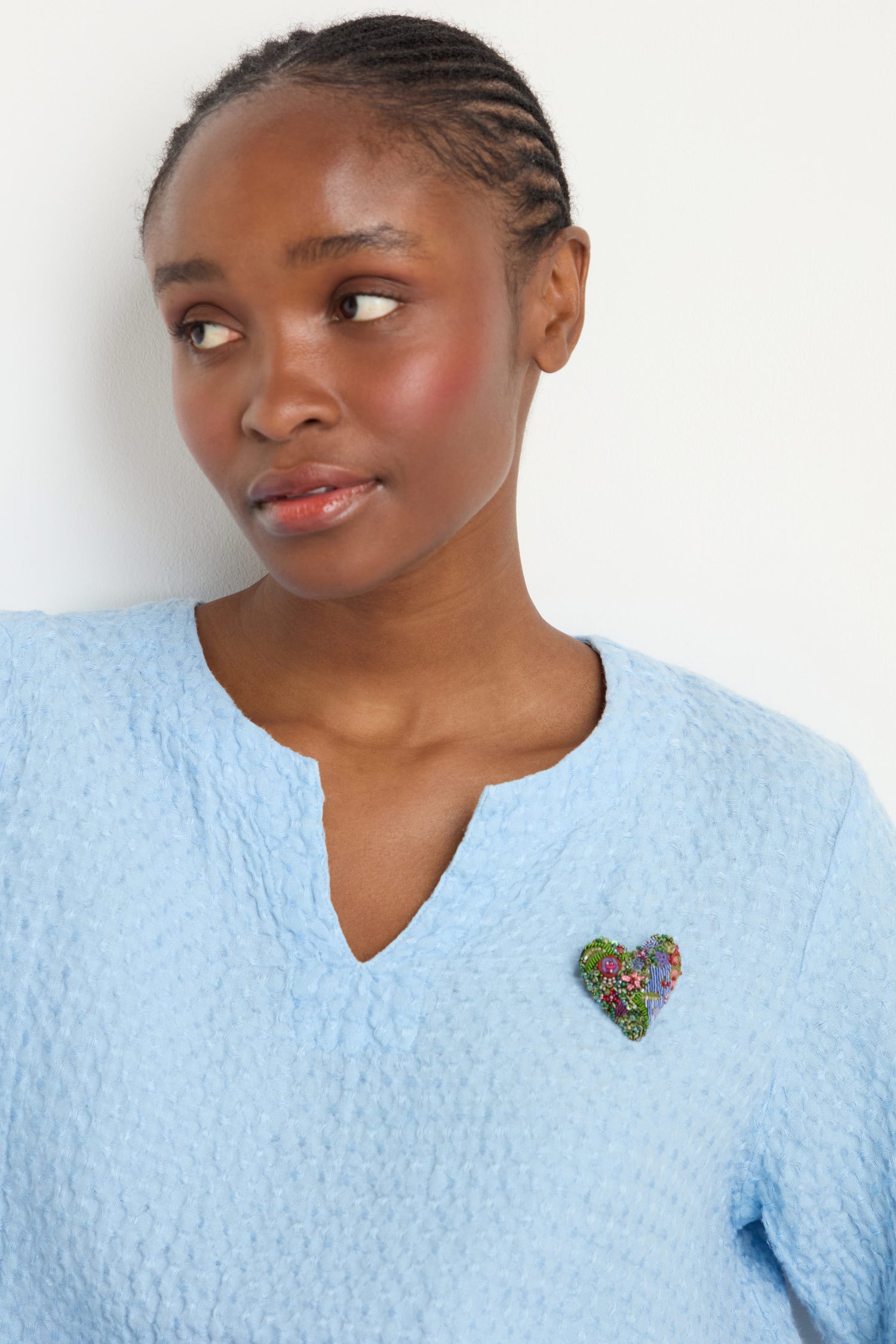 A woman in a light blue textured V-neck top looks to the side, wearing the Hand Embroidered Blooming Heart Brooch on her chest for a colorful, beaded accent.