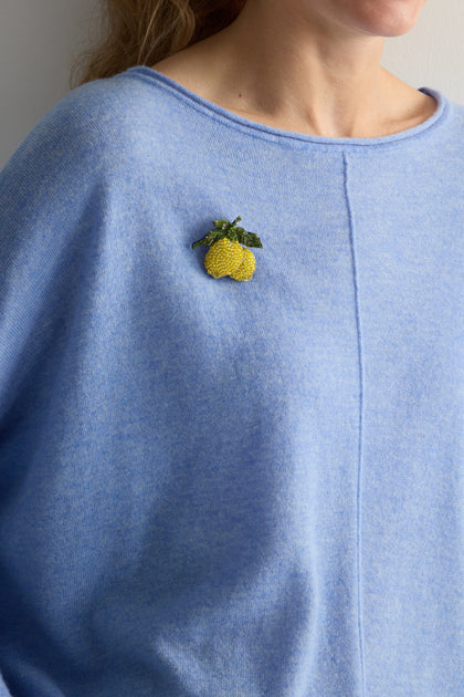 A person wearing a light blue sweater featuring a Hand Beaded Amalfi Lemons Brooch, capturing the essence of Mediterranean beauty.