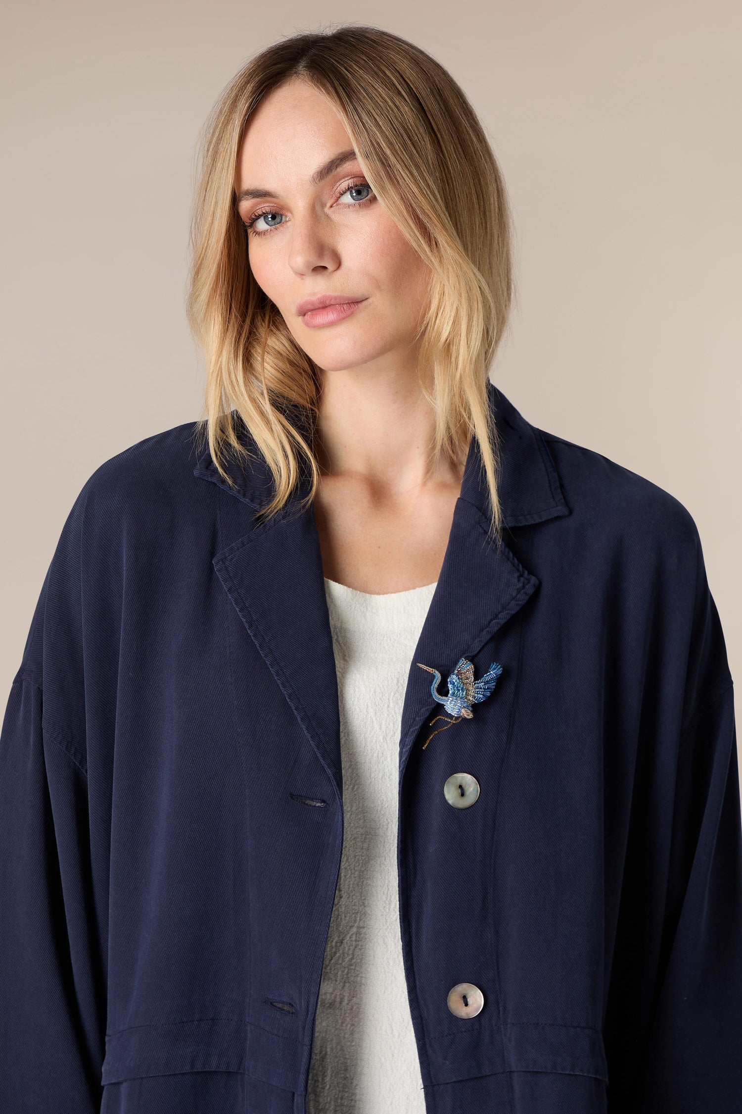 A woman wearing a navy jacket and white shirt accessorized with a Hand Embroidered Blue Heron Brooch.