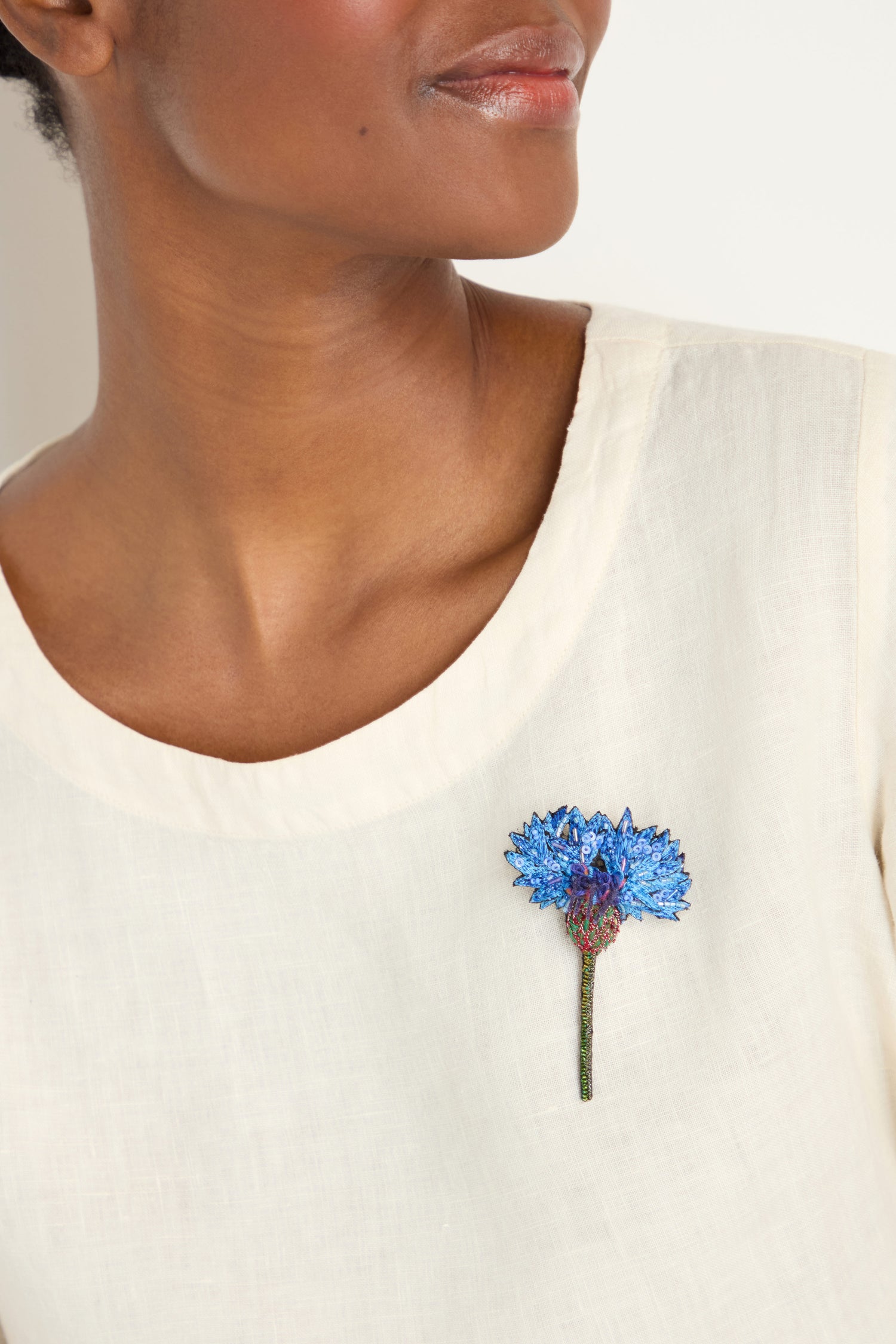 A person wears a cream-colored top adorned with the Hand Embroidered Blue Cornflower Brooch, a beautiful piece of botanical jewelry, pinned to the chest.