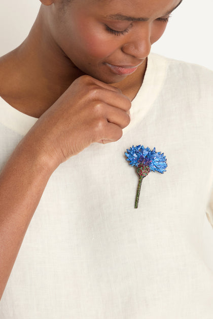 A person wearing a white shirt holds their hand near a Hand Embroidered Blue Cornflower Brooch, a striking piece of botanical jewelry, pinned to their chest.