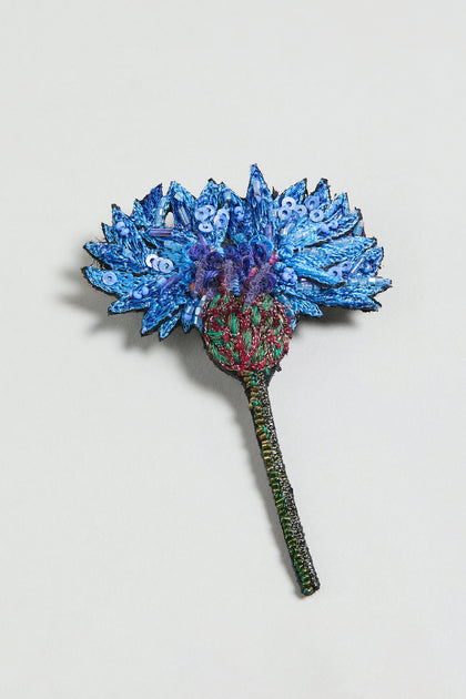 The Hand Embroidered Blue Cornflower Brooch showcases detailed blue petals, a red center, and a green stem—an elegant botanical accessory presented on a plain light background.