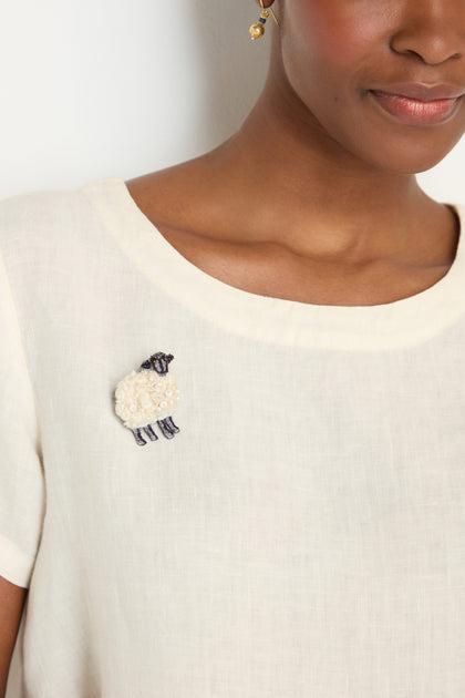 A person wears an off-white top adorned near the neckline with the Hand Embroidered Sheep Brooch.