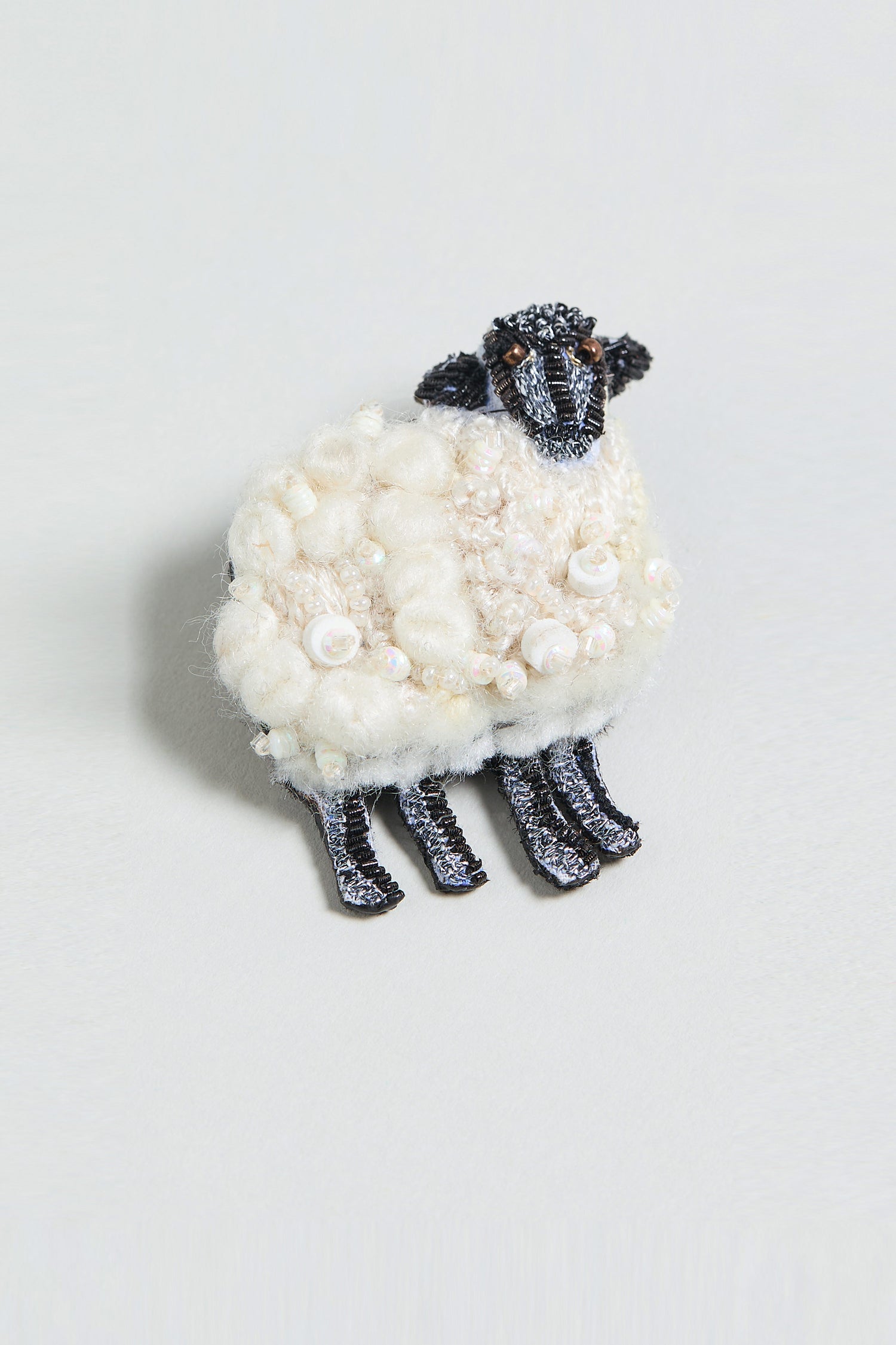 The Hand Embroidered Sheep Brooch features a textured white wool body with a black head and legs, shaped like a sheep, and is beautifully presented on a light background.