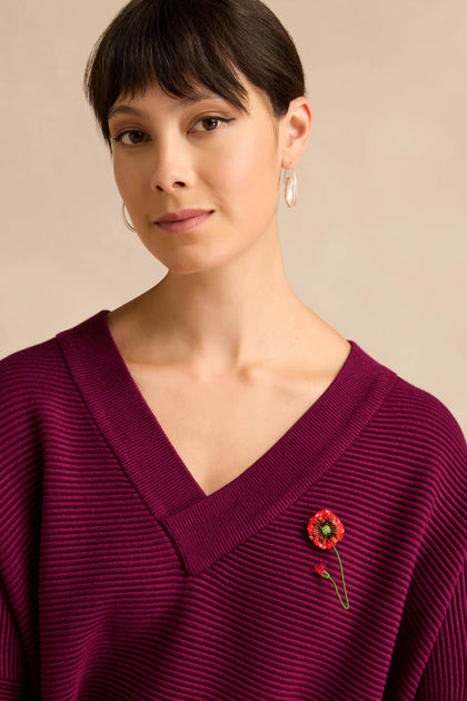 A woman with short brown hair wears a deep purple V-neck sweater and hoop earrings, with a Hand Beaded Shirley Poppy Brooch pinned to her sweater.