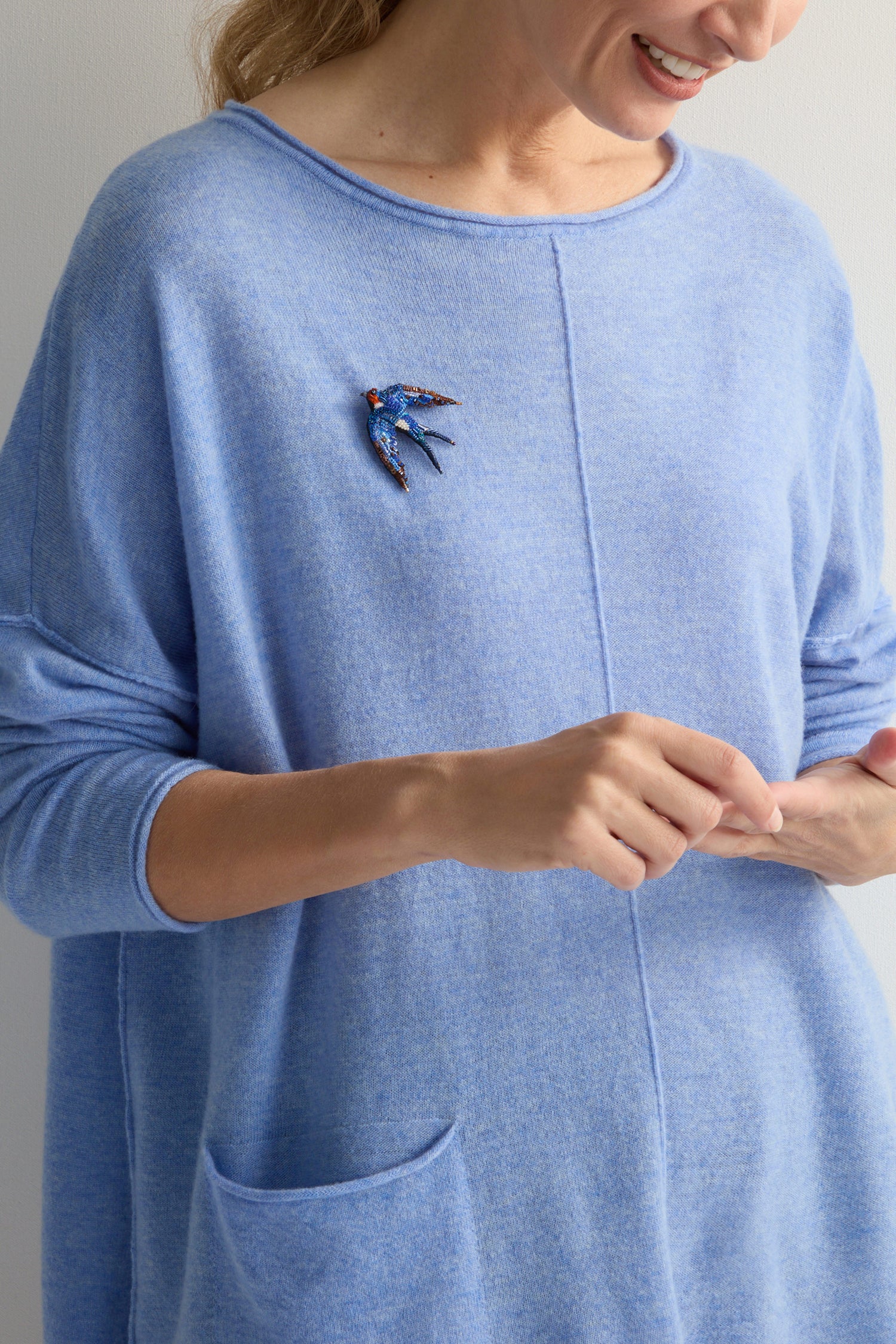 Against a plain background, a person in a light blue sweater wears the Hand Beaded Flying Swallow Brooch. The intricate beadwork of the bird-shaped accessory sparkles elegantly, showcasing its detailed craftsmanship.