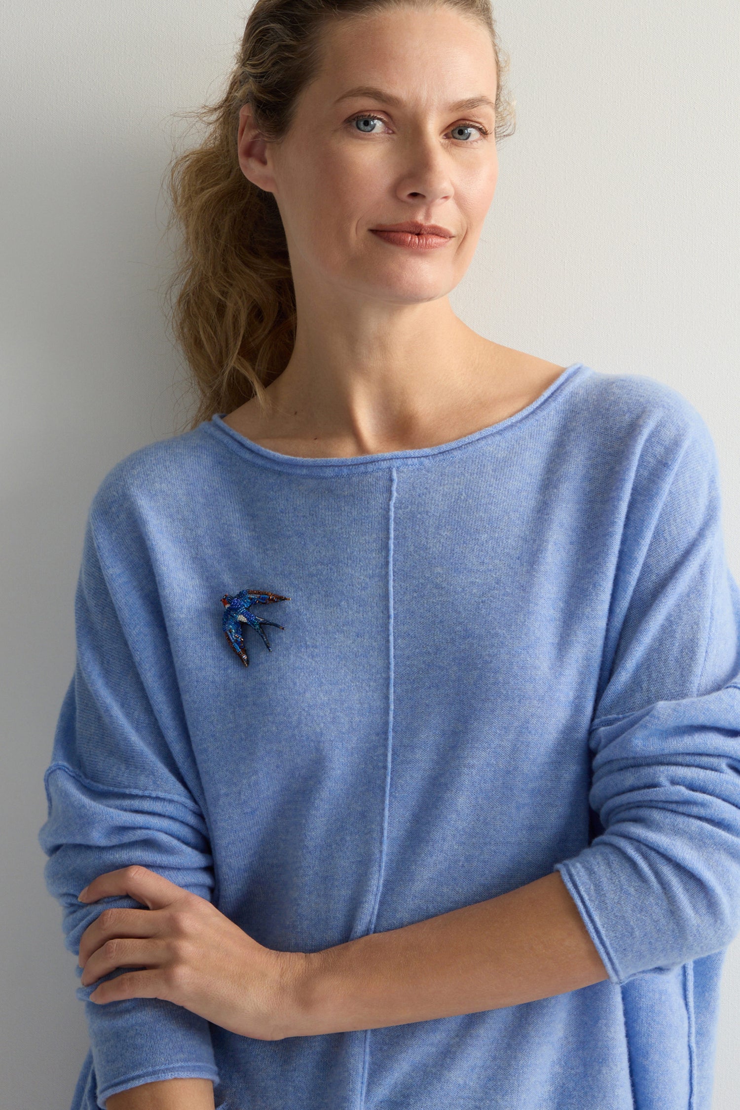 A person stands against a white background wearing a light blue sweater with a Hand Beaded Flying Swallow Brooch on the shoulder, facing forward.