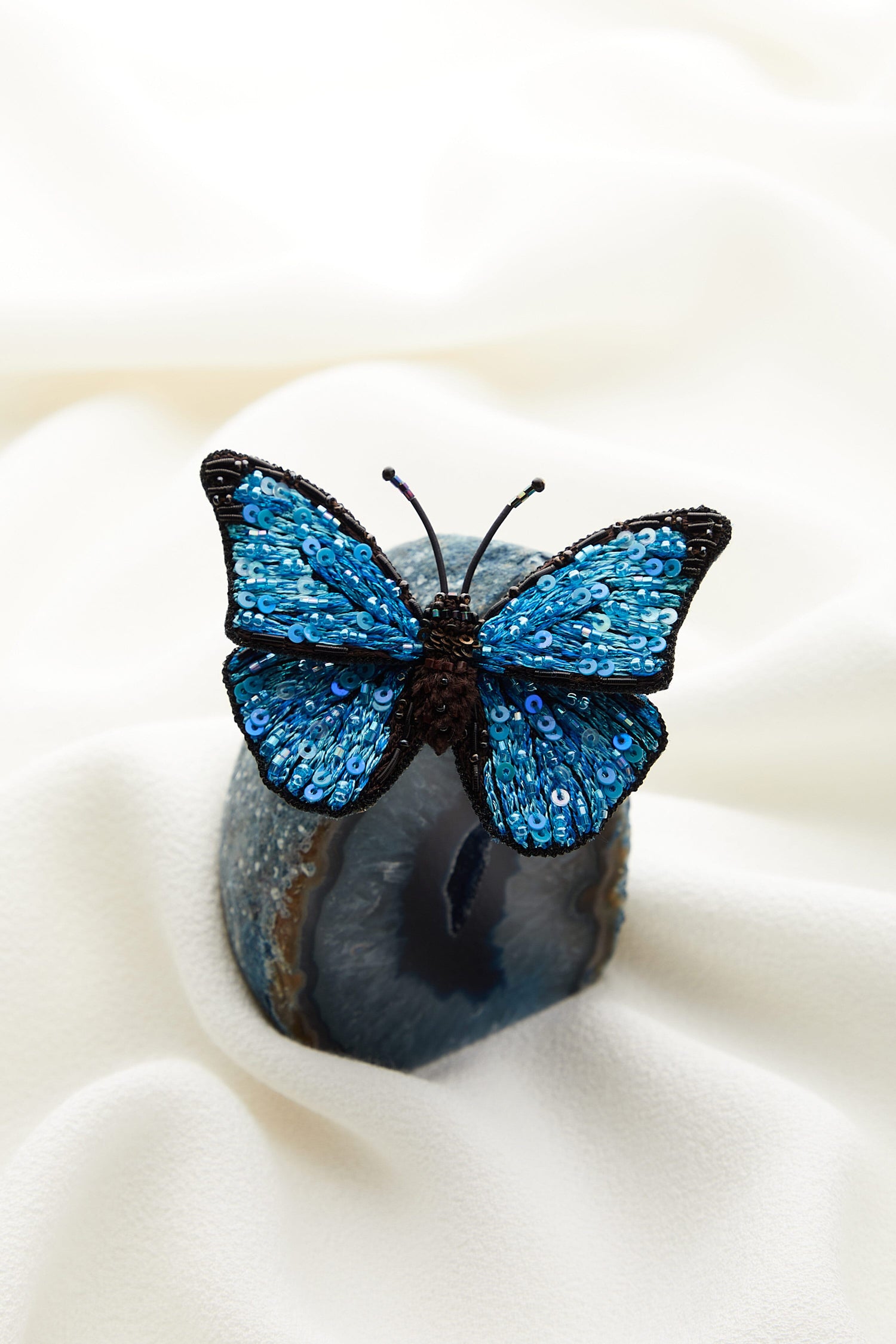 A Hand Embroidered Morpho Butterfly Brooch, hand-beaded with intricate details, rests gracefully on top of a piece of cloth.