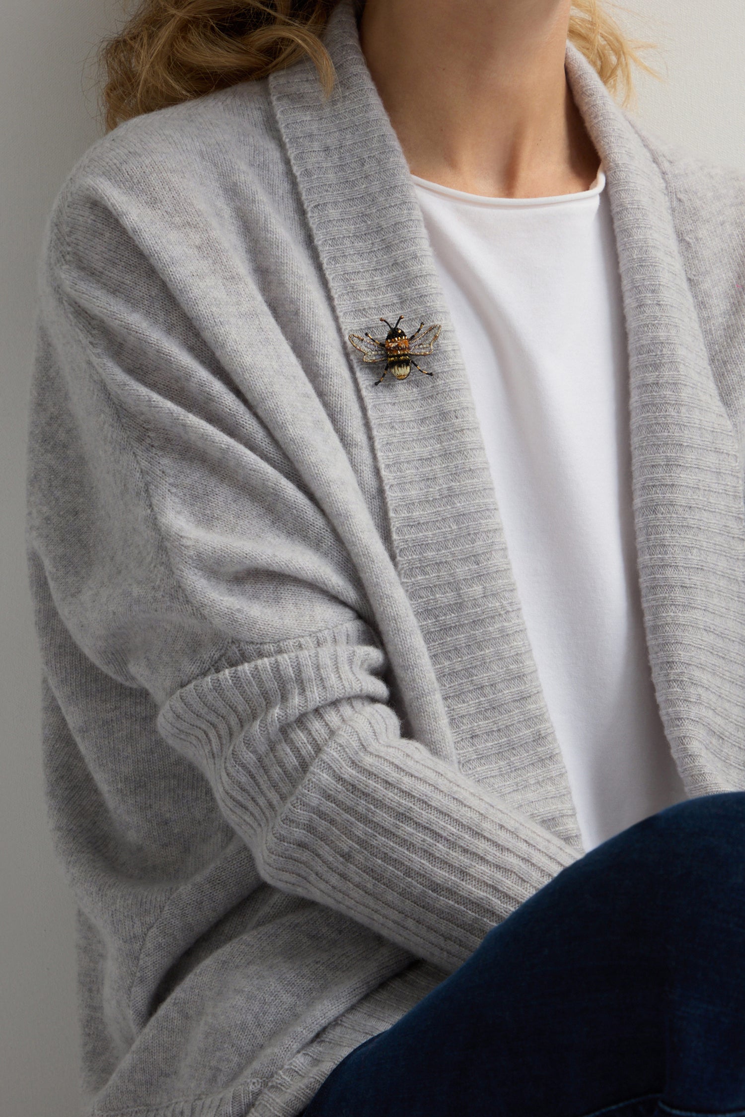 A person, dressed in a gray cardigan and white shirt, adorns their outfit with the elegant Hand Beaded Humble Bee Brooch, adding a touch of sustainable sophistication.