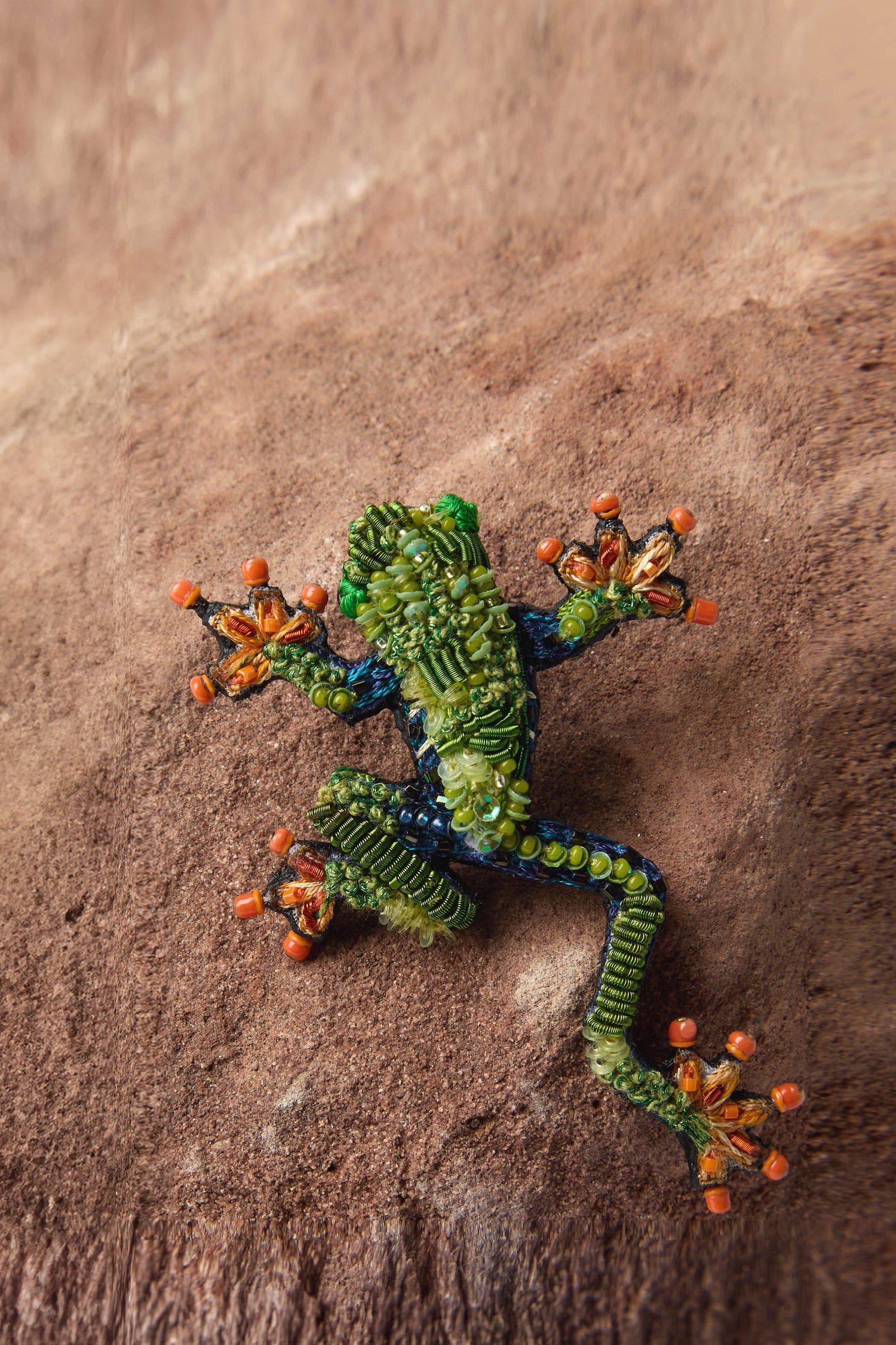 The Hand Beaded Tree Frog Brooch by Trovelore features a green body and vibrant orange toes, crafted with intricate detail, displayed on a brown textured surface.