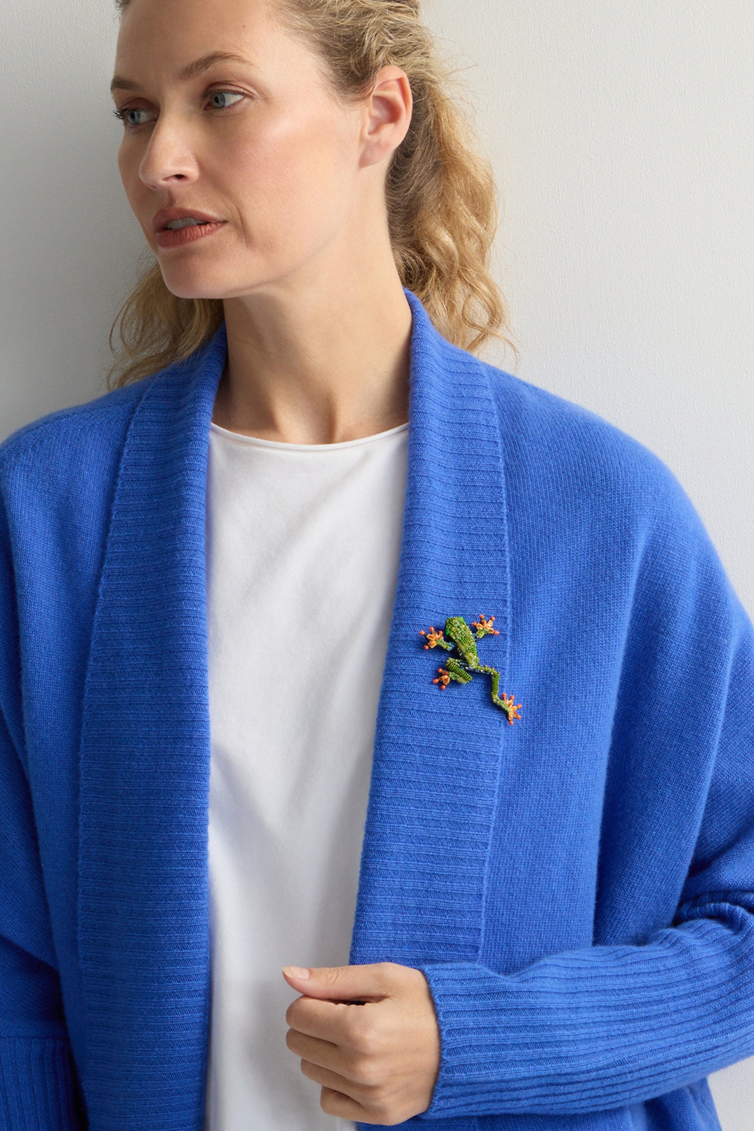 A woman in a blue cardigan wears Trovelore's Hand Beaded Tree Frog Brooch, standing against a plain background.